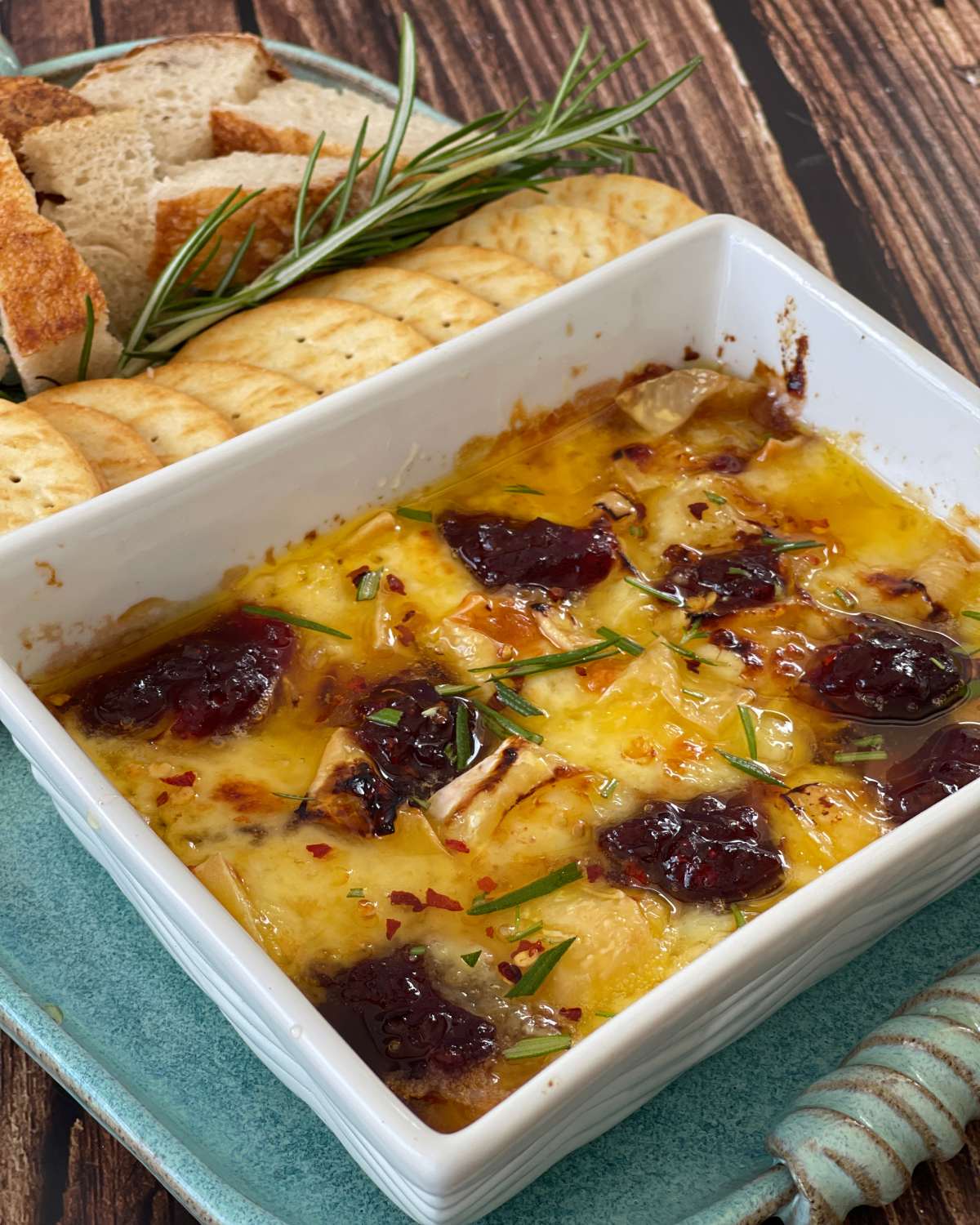 Air Fryer Cranberry Brie Dip Baked brie in a square dish topped with cranberry sauce and rosemary, served with sliced bread and crackers.