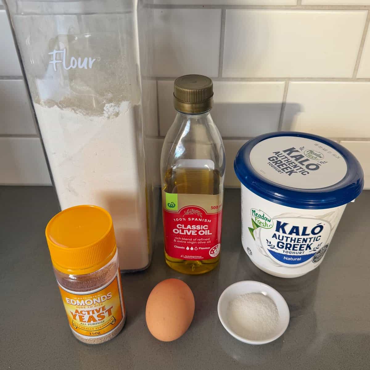 Homemade Garlic Naan Ingredients for a recipe on a counter: a container of flour, a bottle of olive oil, a tub of Greek yogurt, a jar of active yeast, a raw egg, and a small bowl of sugar.