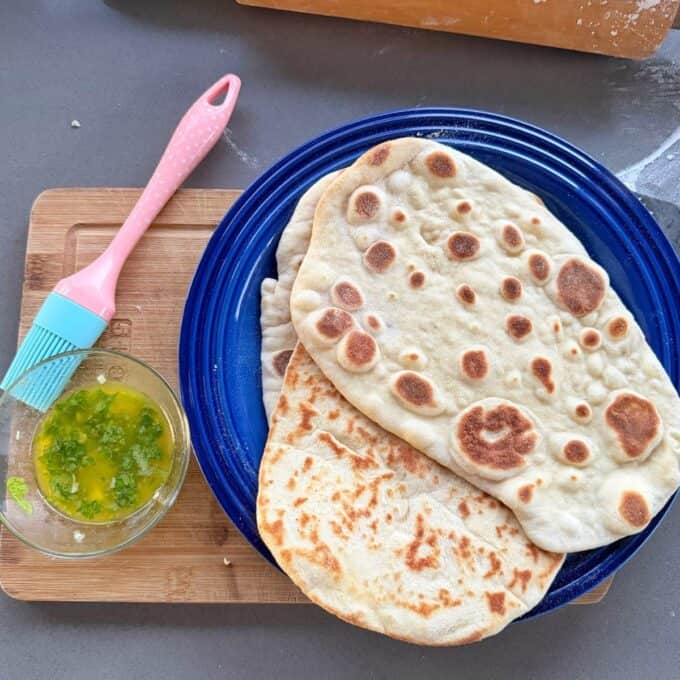 Homemade Garlic Naan Two pieces of flatbread on a blue plate next to a small bowl of herb butter and a silicone pastry brush.