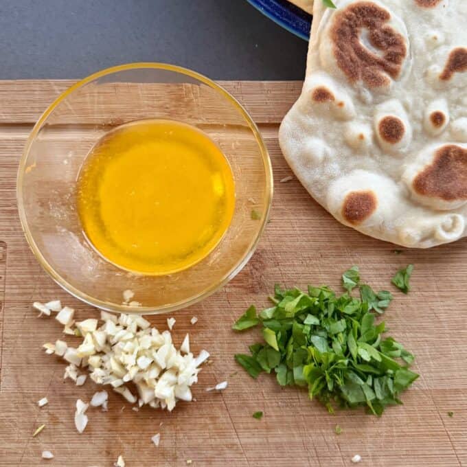 Homemade Garlic Naan A glass bowl of melted butter next to piles of chopped garlic and parsley on a wooden cutting board, with a piece of flatbread nearby.