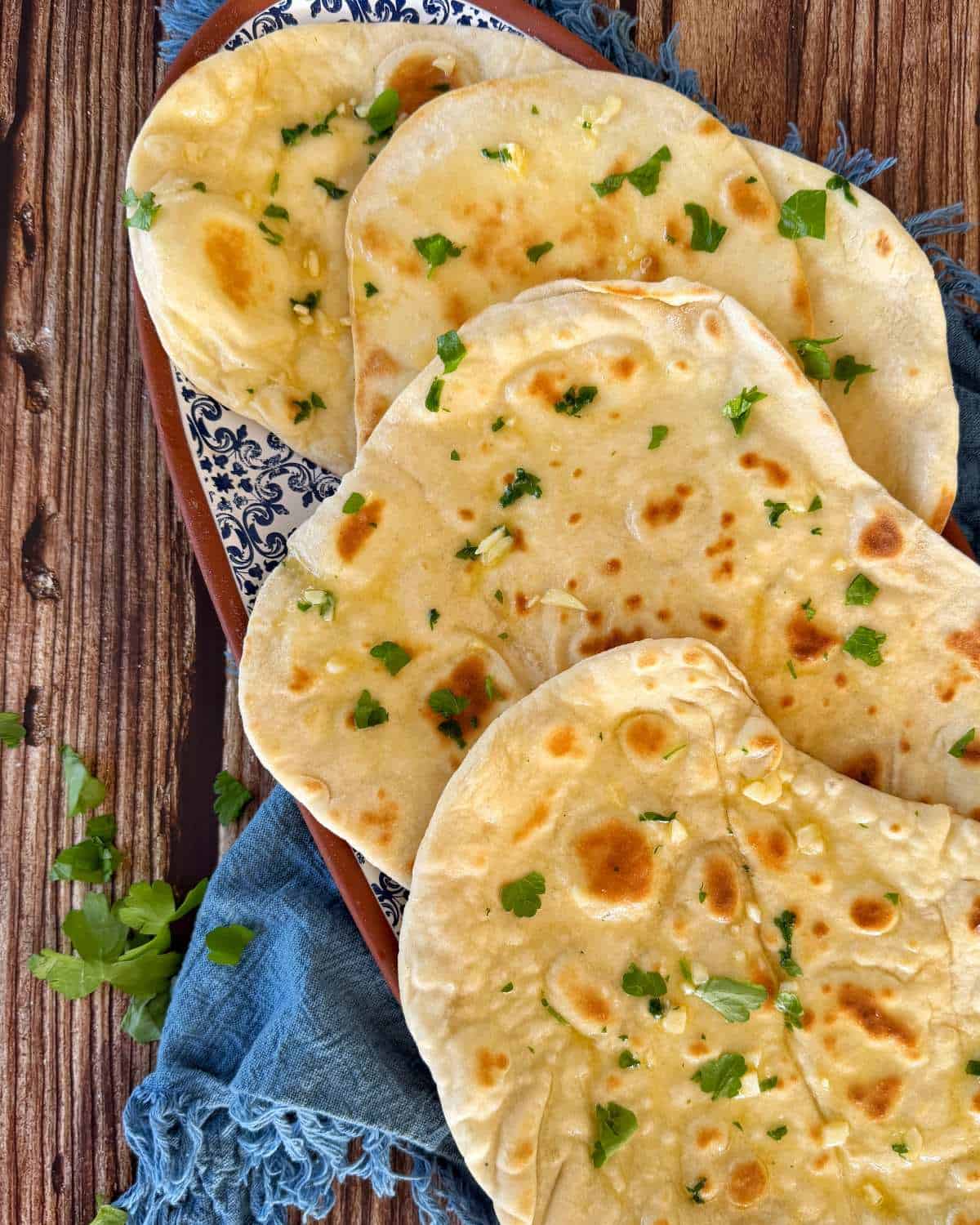 Homemade Garlic Naan A stack of garlic naan bread topped with fresh parsley on a patterned serving platter.