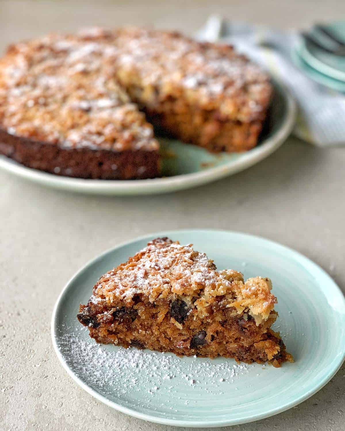Lumberjack Cake A slice of fruit cake with a crumb topping and powdered sugar on a plate, with the rest of the cake in the background.