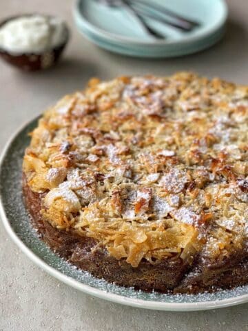 A round cake topped with toasted coconut and a dusting of powdered sugar, served on a green plate with a side of cream.