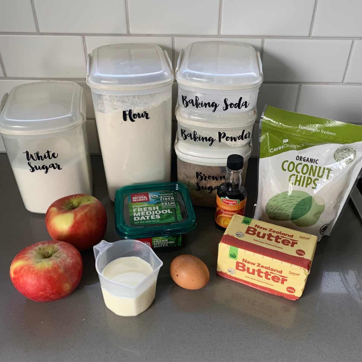 Lumberjack Cake Baking ingredients including two apples, a pitcher of milk, an egg, butter, vanilla extract, coconut chips, dates, and labeled containers of flour, sugar, baking soda, and baking powder.