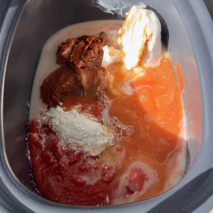 Slow Cooker Butter Chicken A slow cooker bowl containing raw ingredients: tomato sauce, coconut milk, curry paste, dollops of yogurt, and a scoop of flour.