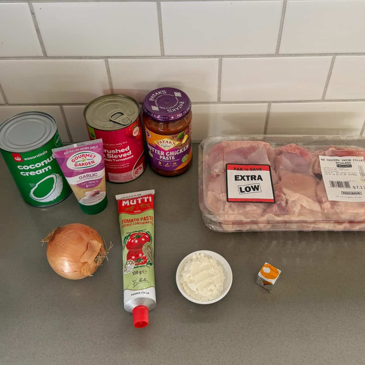 Ingredients for butter chicken: a can of coconut cream, garlic paste, crushed tomatoes, butter chicken paste, raw chicken thighs, an onion, a tube of tomato paste, a small bowl of flour, and a stock cube.