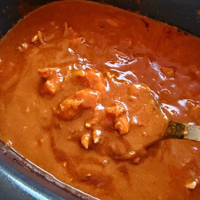 Slow Cooker Butter Chicken A metal spoon lifting chicken pieces from a thick, reddish-brown curry in a slow cooker.