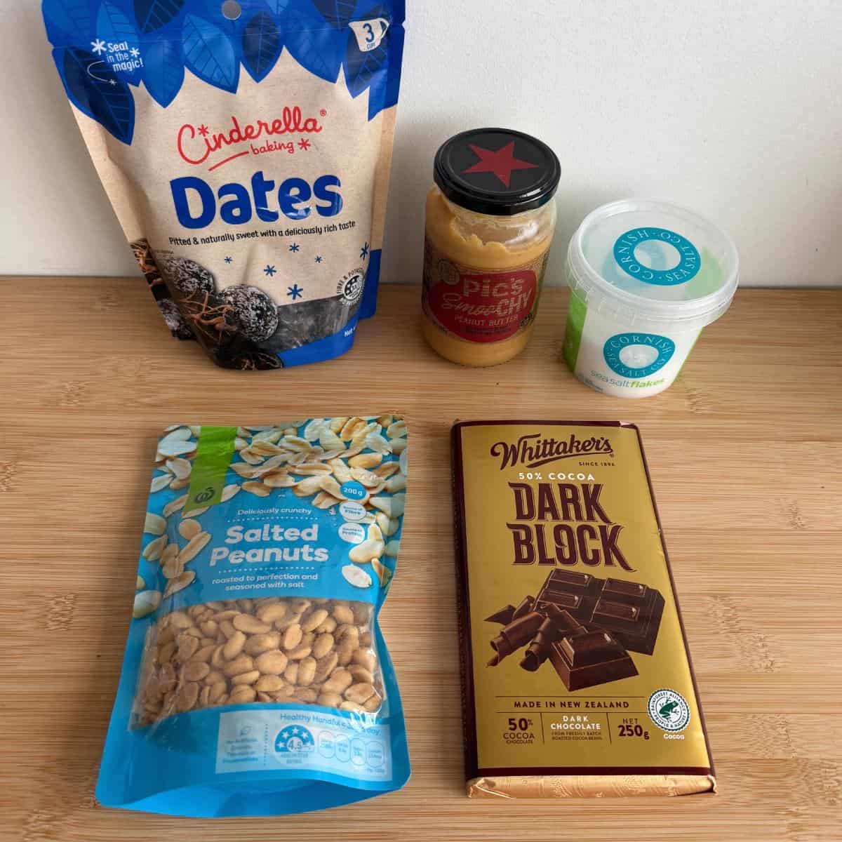 Snickers Peanut Butter Bites A bag of Cinderella pitted dates, a jar of Pics peanut butter, a tub of Cornish sea salt flakes, a bag of salted peanuts, and a bar of Whittakers 50% cocoa dark chocolate on a wooden surface.