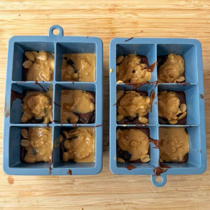 Snickers Peanut Butter Bites Two blue silicone ice cube trays filled with layers of dark chocolate and peanut butter topped with peanuts.