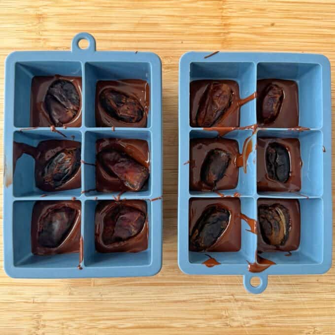 Snickers Peanut Butter Bites Two blue silicone ice cube trays filled with melted chocolate and pitted dates, resting on a wooden surface.