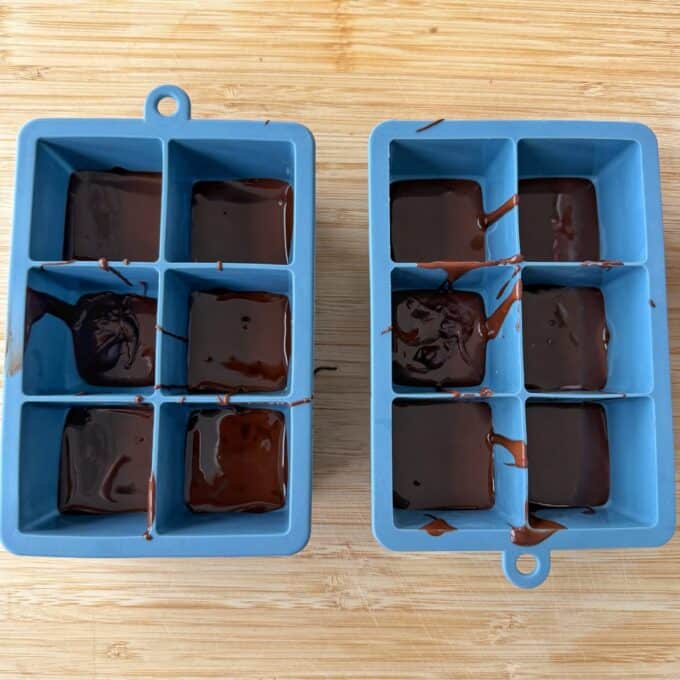 Snickers Peanut Butter Bites Two blue silicone ice cube trays filled with melted chocolate, resting on a wooden surface.